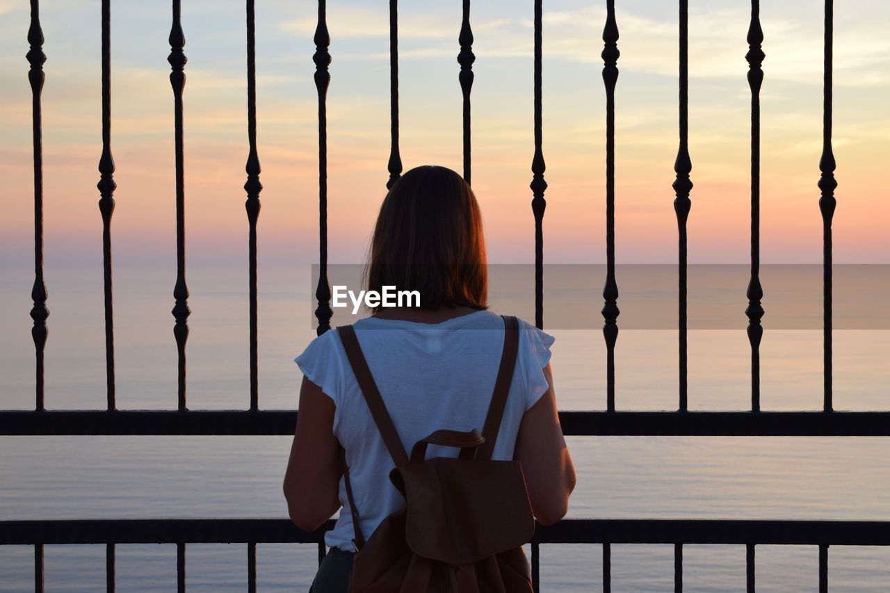 Rear view of woman looking at sea through railing against sky during sunset