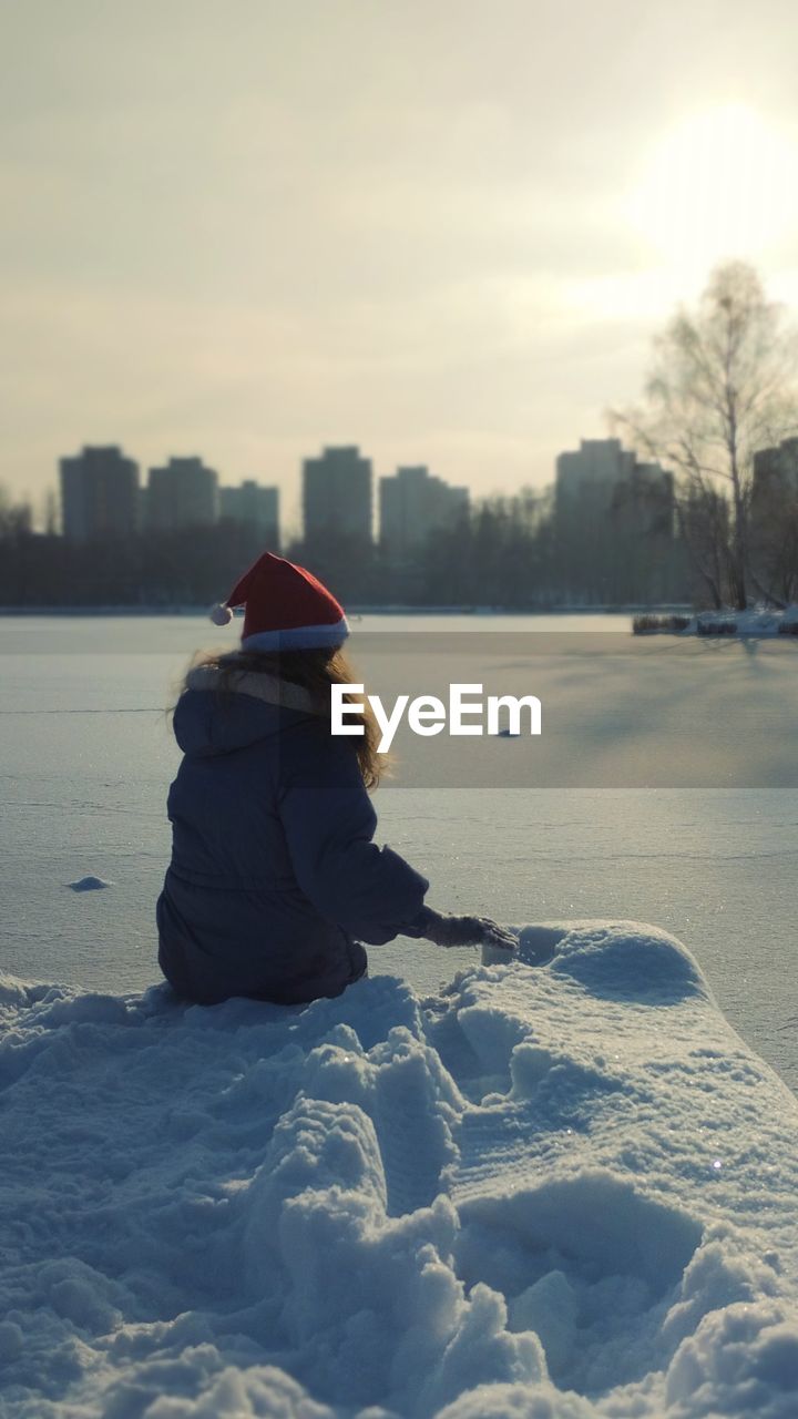 winter, cold temperature, one person, snow, nature, clothing, warm clothing, freezing, sky, hat, rear view, women, ice, child, leisure activity, childhood, knit hat, blue, landscape, architecture, water, day, sitting, morning, environment, full length, sunlight, outdoors, adult, frozen, female, lifestyles, beauty in nature, land, cloud, reflection, city, building exterior, holiday, built structure
