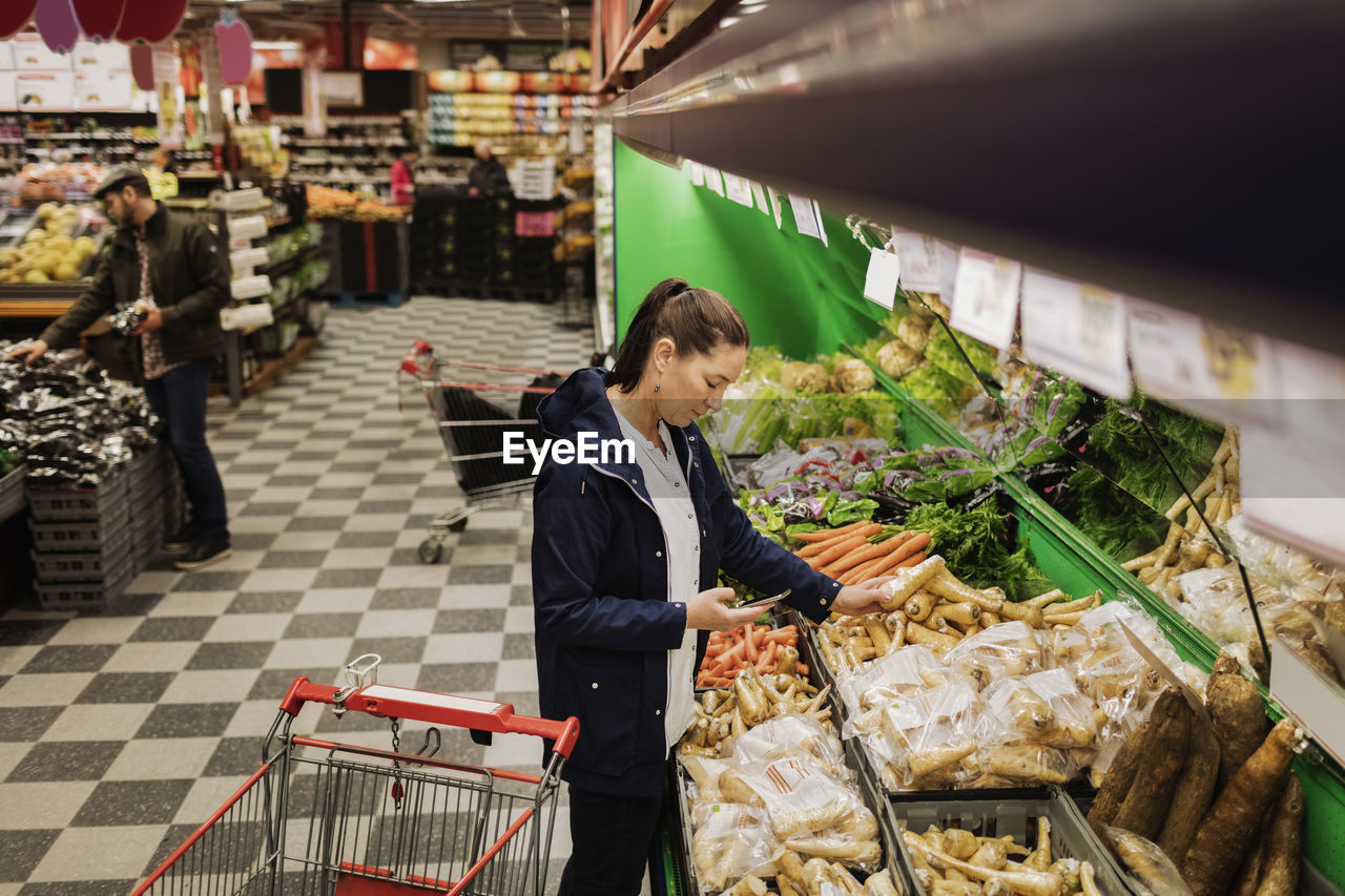 High angle view of woman holding phone while buying vegetables in supermarket