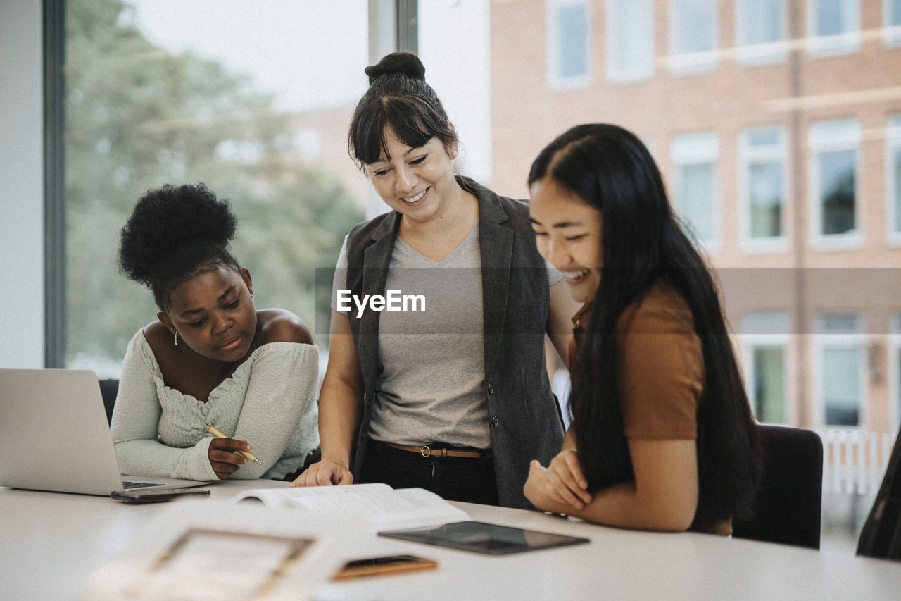 Smiling female professor explaining multiracial students at table in university