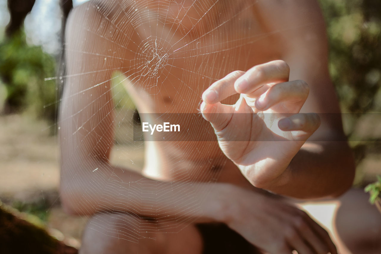 Midsection of shirtless man holding spider seen through web