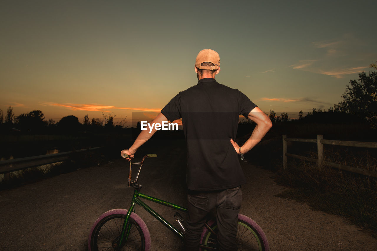 Rear view of young man with bicycle standing on road against sky during sunset