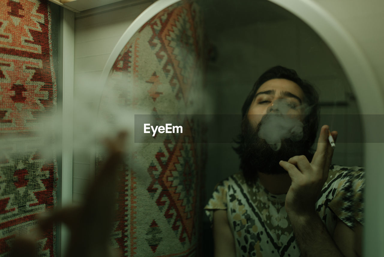 Bearded man smoking cigarette reflecting on mirror at home