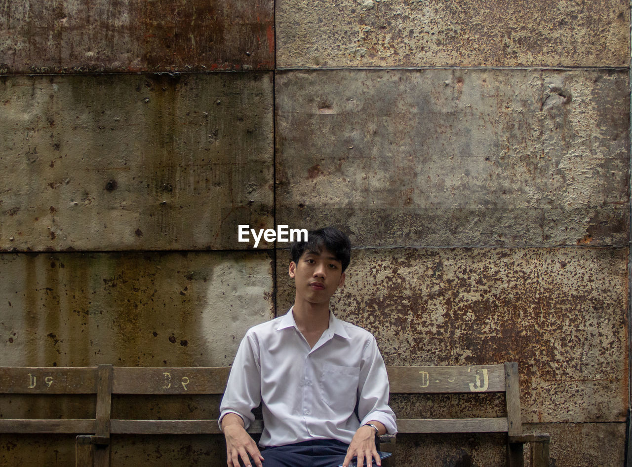 Portrait of young man sitting on bench against wall