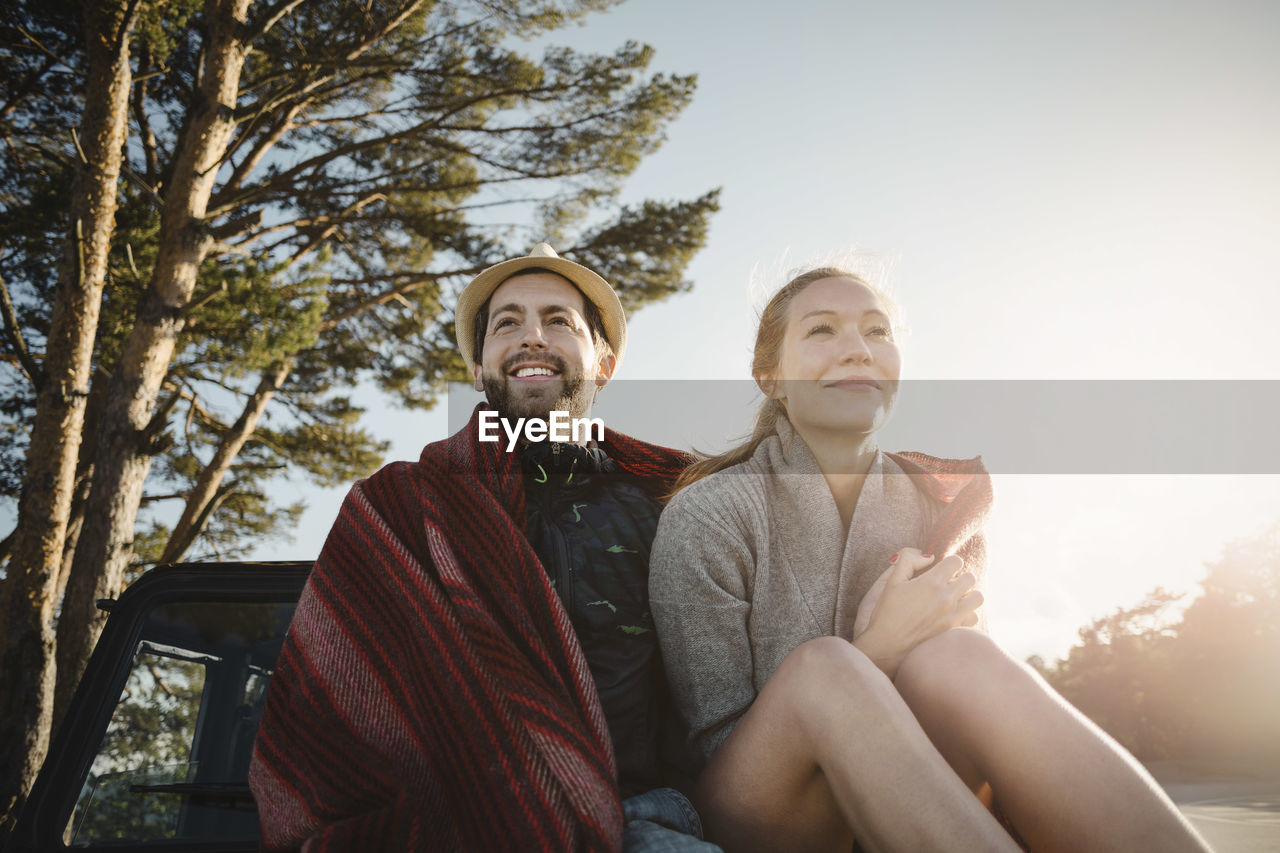 Happy wonderlust couple wrapped in blanket sitting on jeep against clear sky