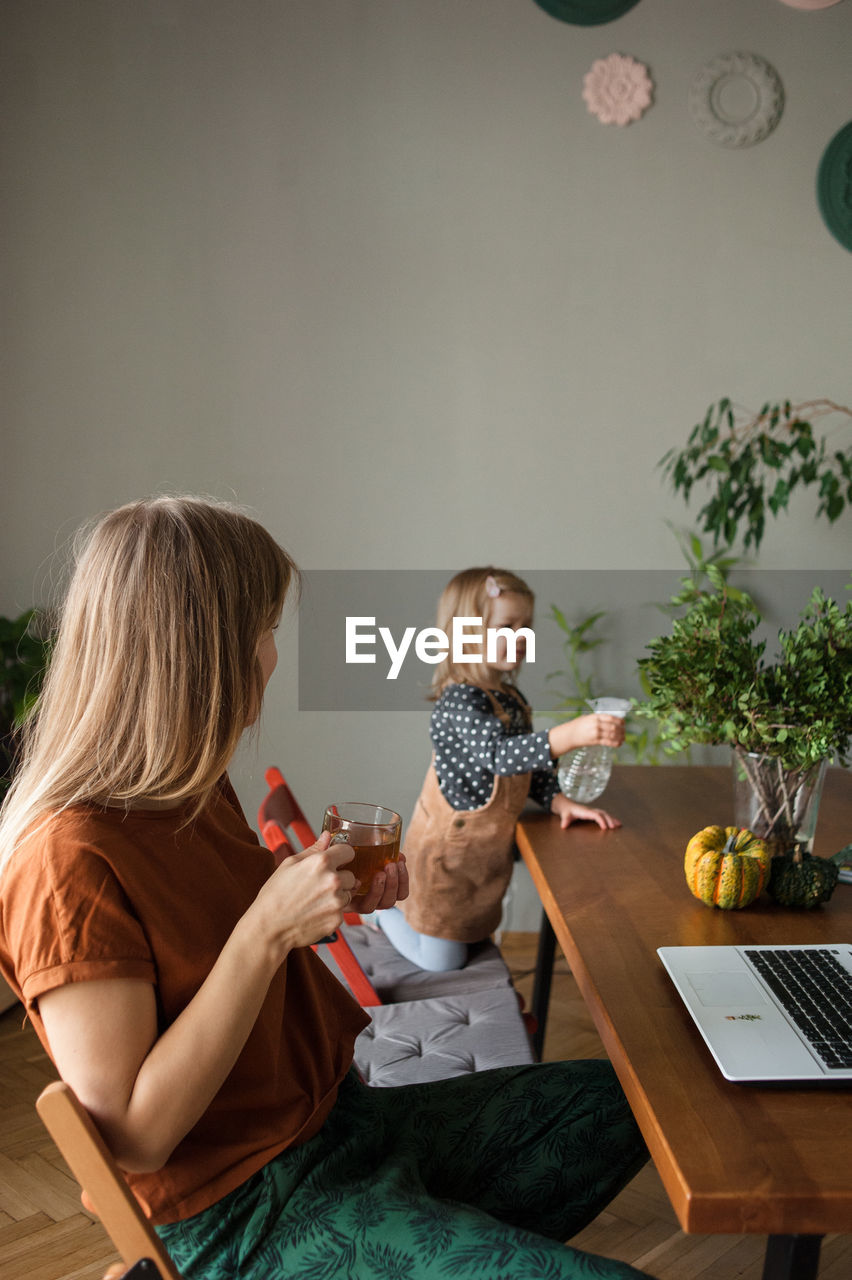 Mother with tea cup rests and looks at child who watering green plants