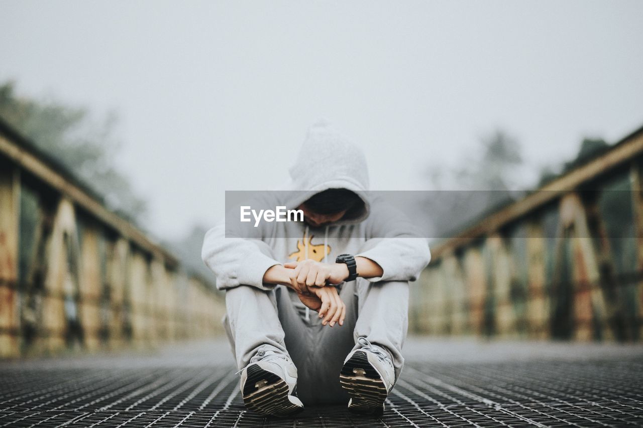 Man wearing hooded shirt while sitting on bridge