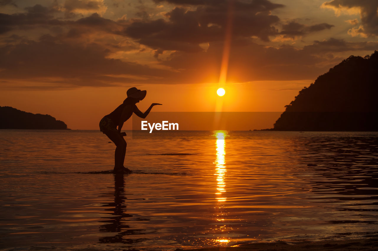 Silhouette woman blowing kiss while standing in sea against sky during sunset