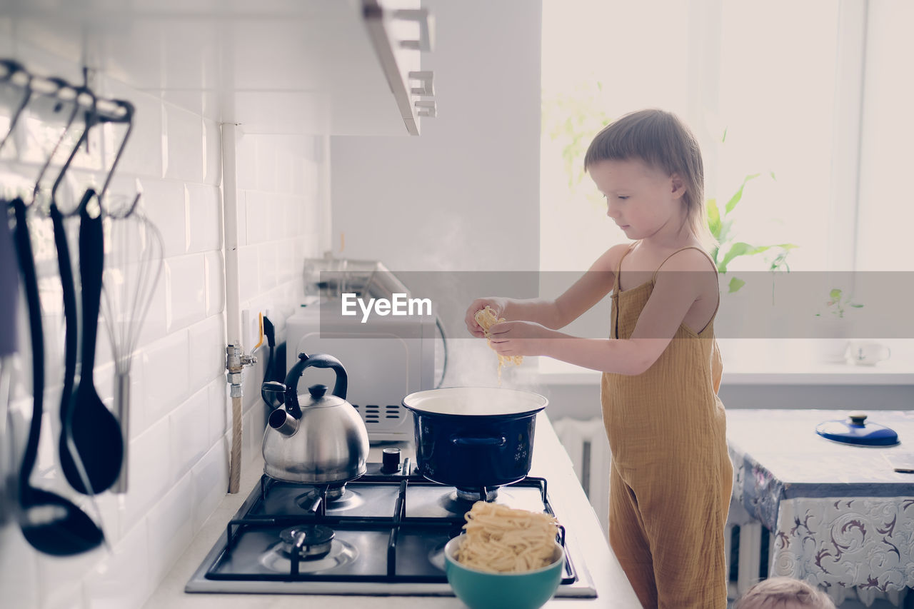 Cute toddler child in kitchen by stove helps mom cook, bright interior of kitchen