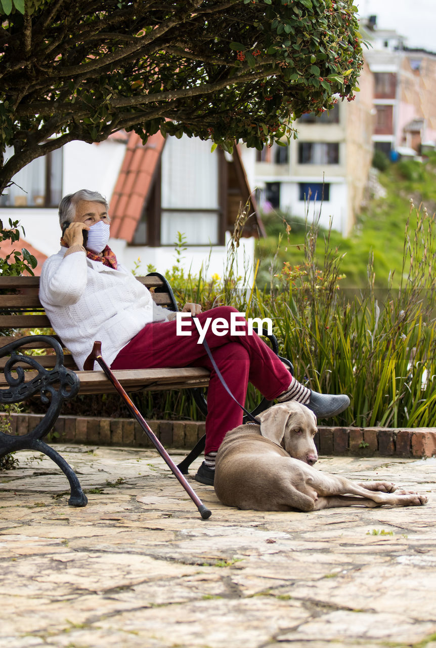 Full length of senior wearing mask with dog sitting outdoors