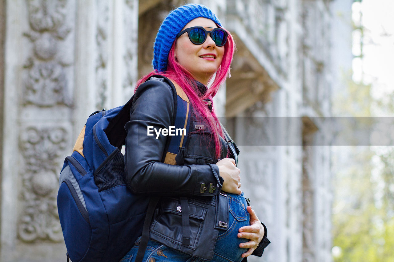 one person, adult, clothing, fashion, architecture, blue, sunglasses, women, spring, glasses, person, smiling, backpack, happiness, young adult, jacket, lifestyles, leisure activity, city, standing, day, outdoors, luggage and bags, emotion, nature, portrait, human face, warm clothing, hat, side view, travel, waist up, focus on foreground, winter, scarf, cheerful, copy space, casual clothing, travel destinations, female, enjoyment, arts culture and entertainment, tourism, looking, city life, holiday, leather jacket, three quarter length, goggles, trip, fashion accessory, vacation, tourist
