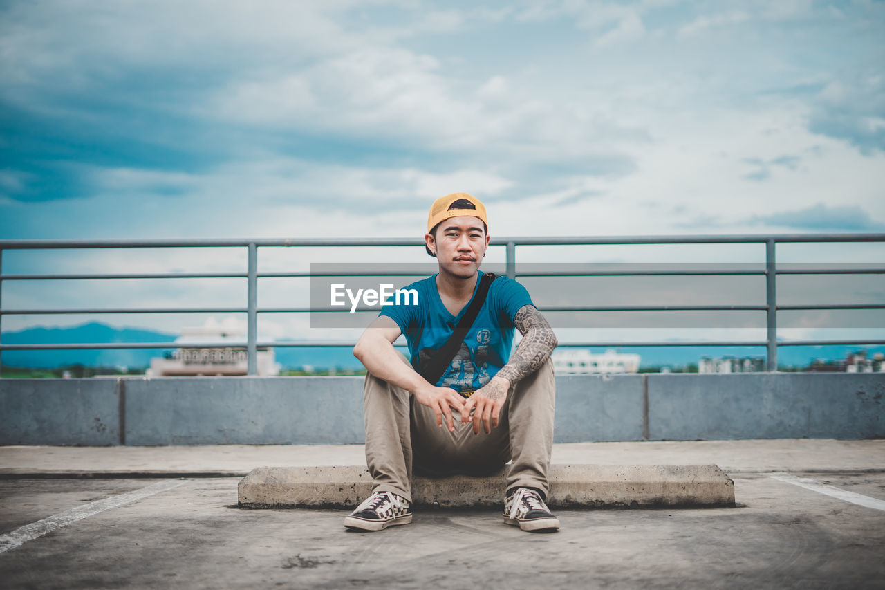 Portrait of young man sitting on street in city against sky