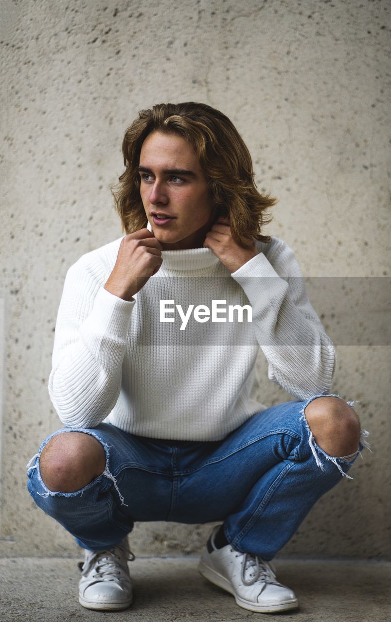 Teenage boy looking away while crouching against wall