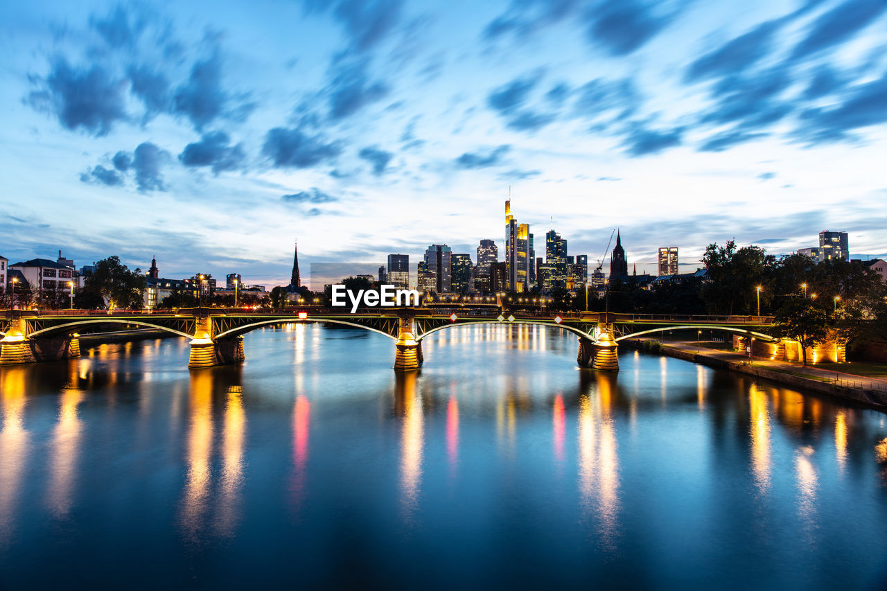 ILLUMINATED BRIDGE OVER RIVER BY BUILDINGS AGAINST SKY