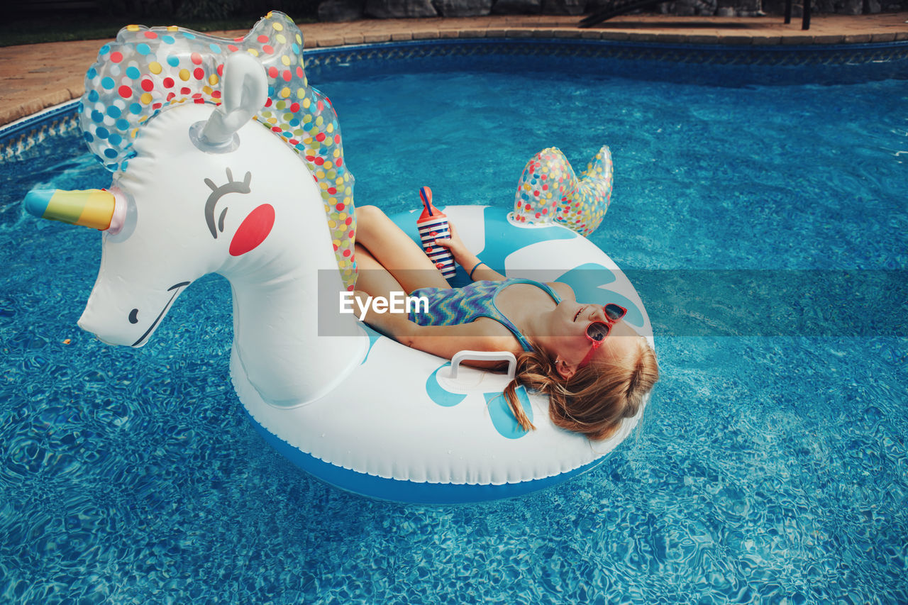 Cute adorable girl in sunglasses with drink lying on inflatable ring unicorn. kid child enjoying 