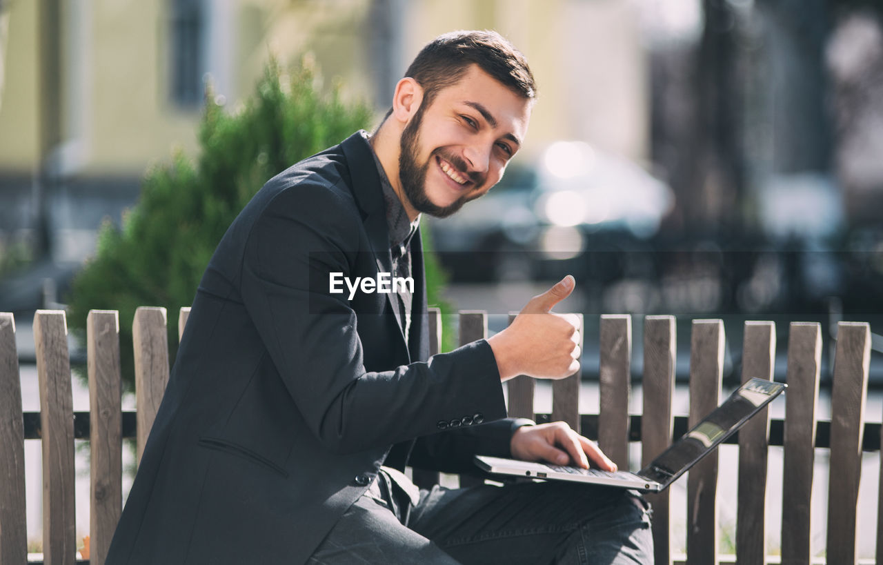 Portrait of smiling businessman gesturing while using laptop by railing