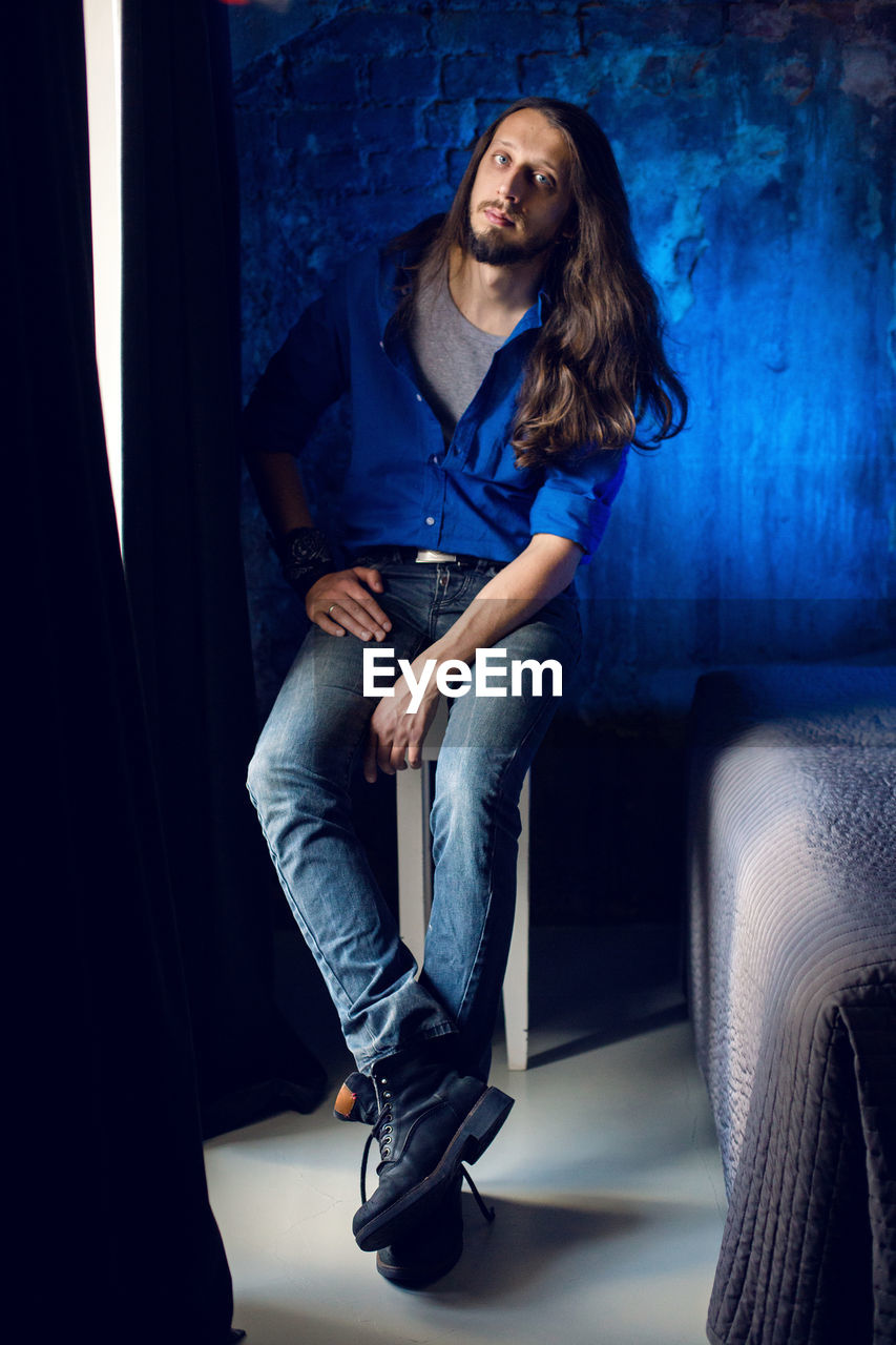 Man with long hair and beard sits at home on a chair by the window in a blue shirt and jeans