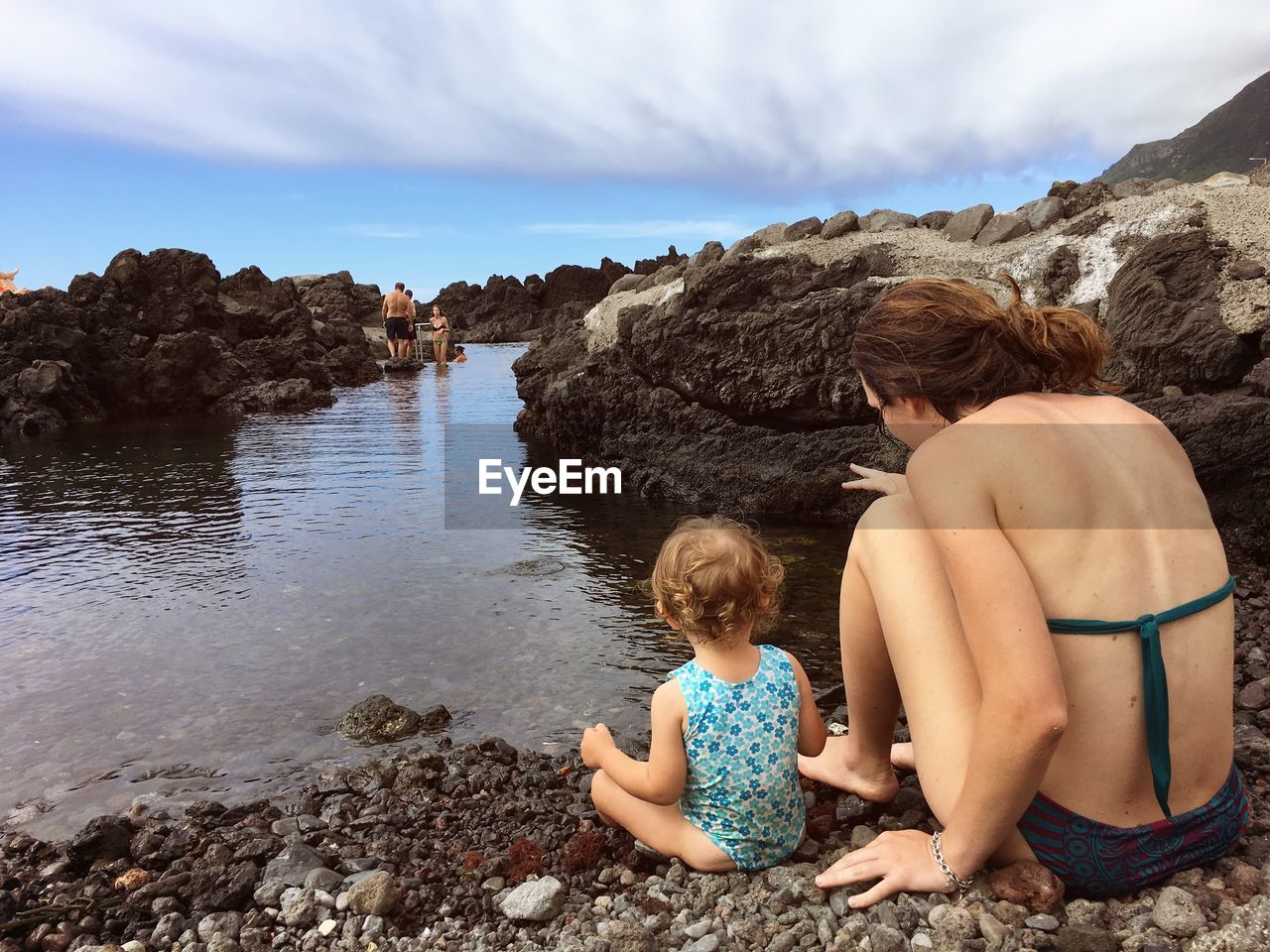 REAR VIEW OF SHIRTLESS BOY SITTING ON ROCK AT BEACH