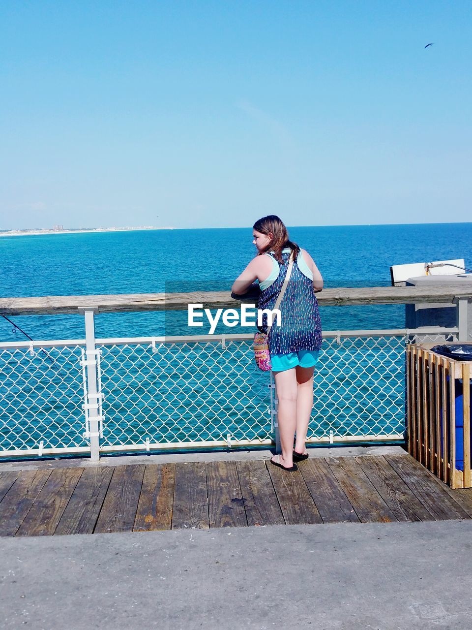 Rear view of woman leaning on railing at pier by sea against clear blue sky