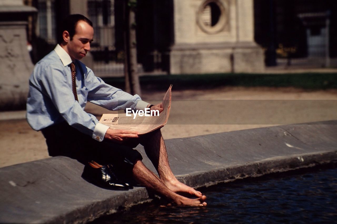 Businessman reading newspaper while sitting by pond in city