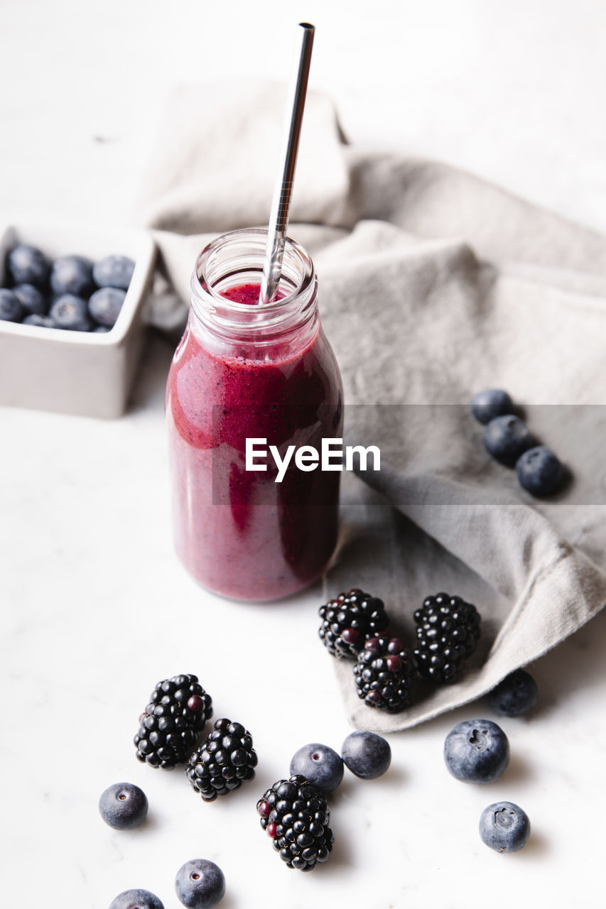 Glass bottle with fresh homemade berry smoothie and a reusable metal straw. some berries near the bottle on a marble surface. high angle vertical image.