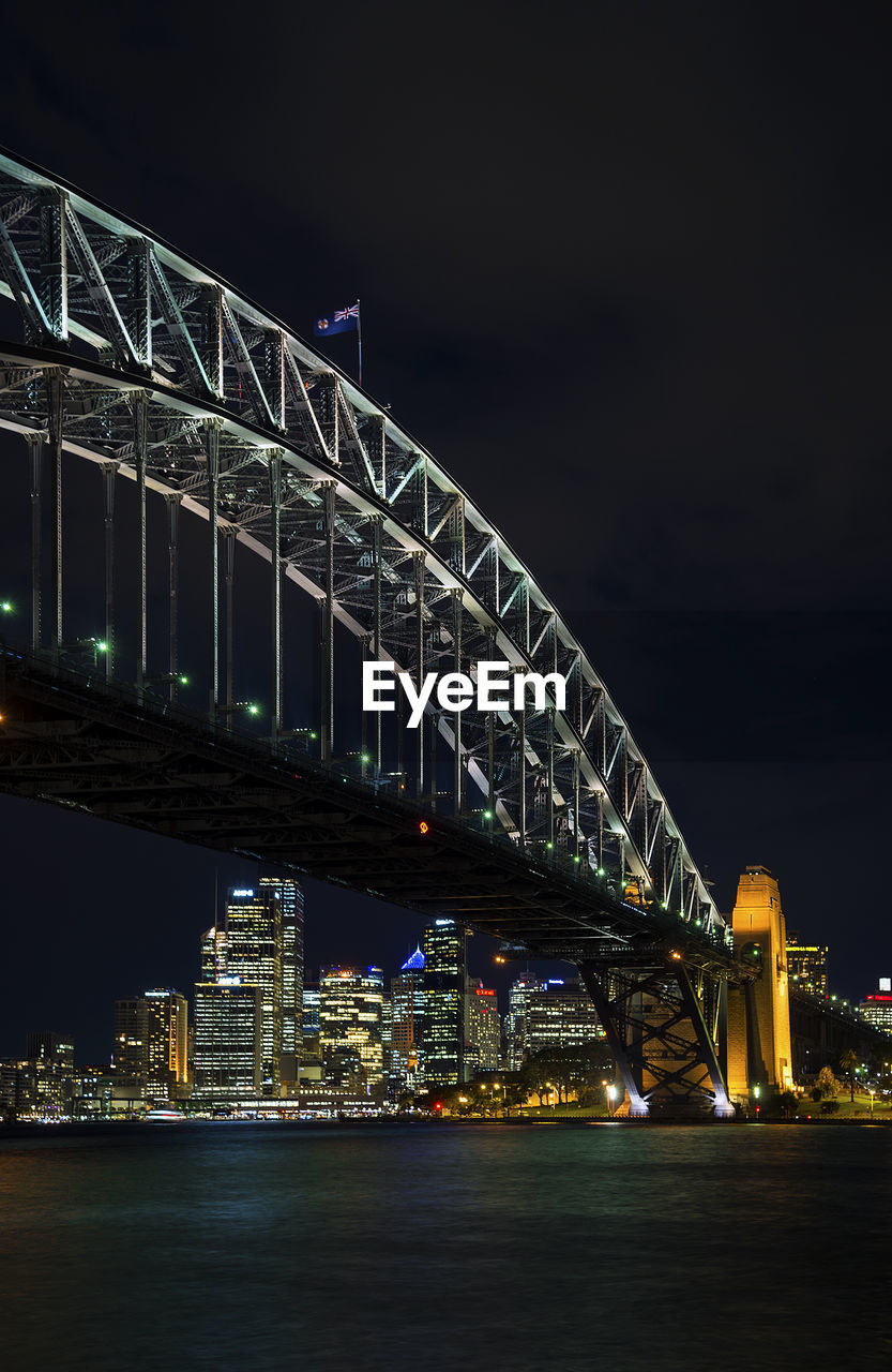 ILLUMINATED BRIDGE OVER RIVER AGAINST BUILDINGS AT NIGHT