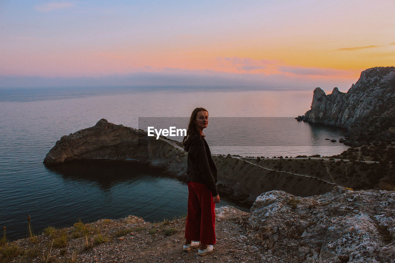 YOUNG WOMAN STANDING ON ROCK AGAINST SEA DURING SUNSET