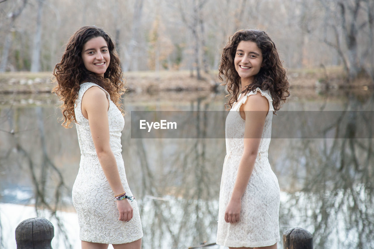 Portrait of smiling sisters wearing white dresses while standing by lake