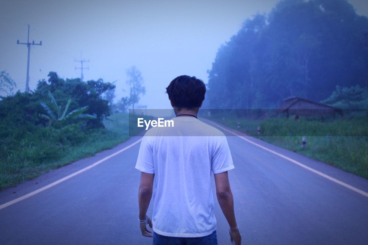 REAR VIEW OF MAN STANDING ON ROAD AMIDST TREES