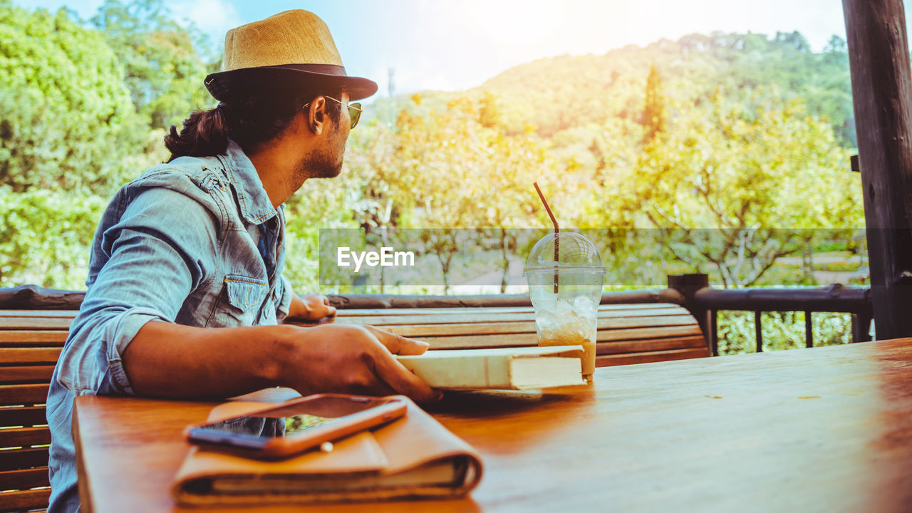 SIDE VIEW OF YOUNG MAN LOOKING AWAY WHILE SITTING ON TABLE AT PARK