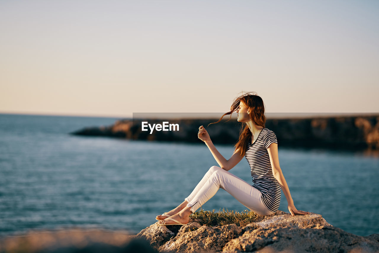 WOMAN SITTING ON ROCK AT SEA SHORE AGAINST SKY