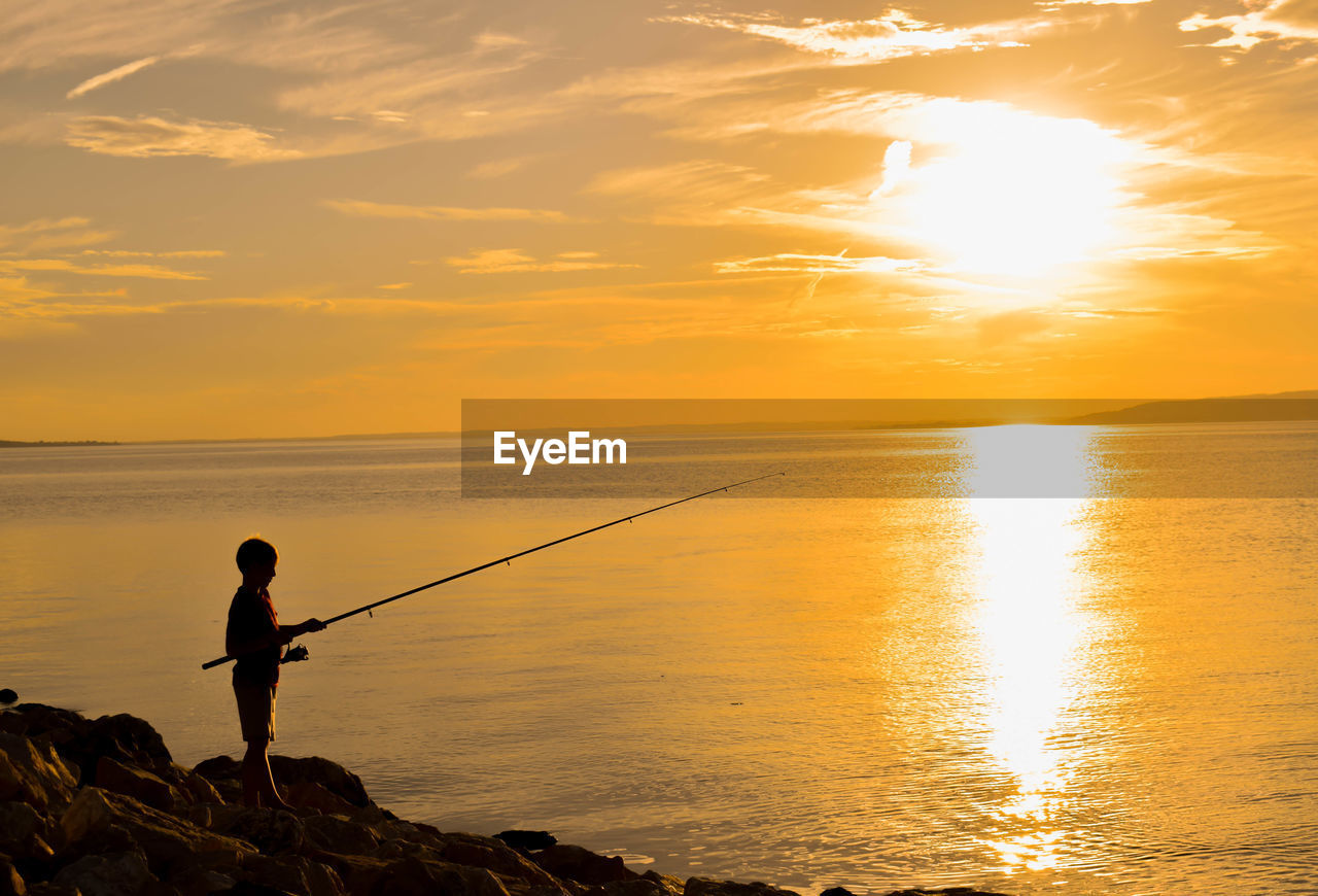 Silhouette boy fishing in sea against sunset sky