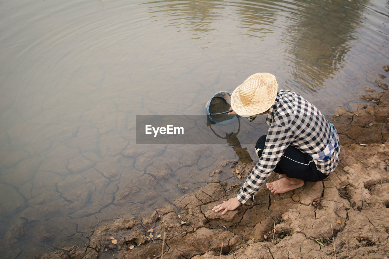 High angle view of person crouching over cracked field while filling water from lake in bucket