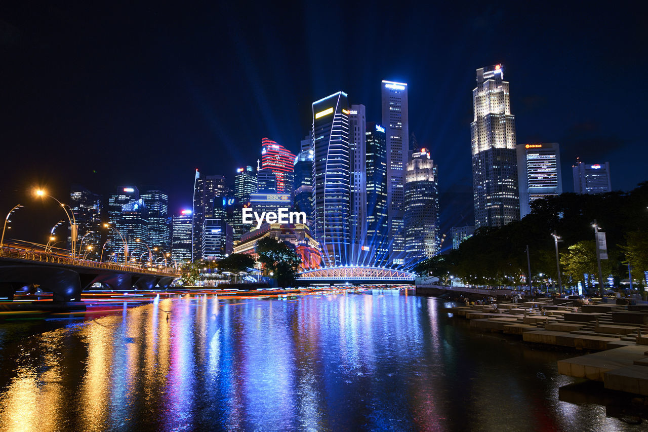 Illuminated bridge over river by buildings against sky at night