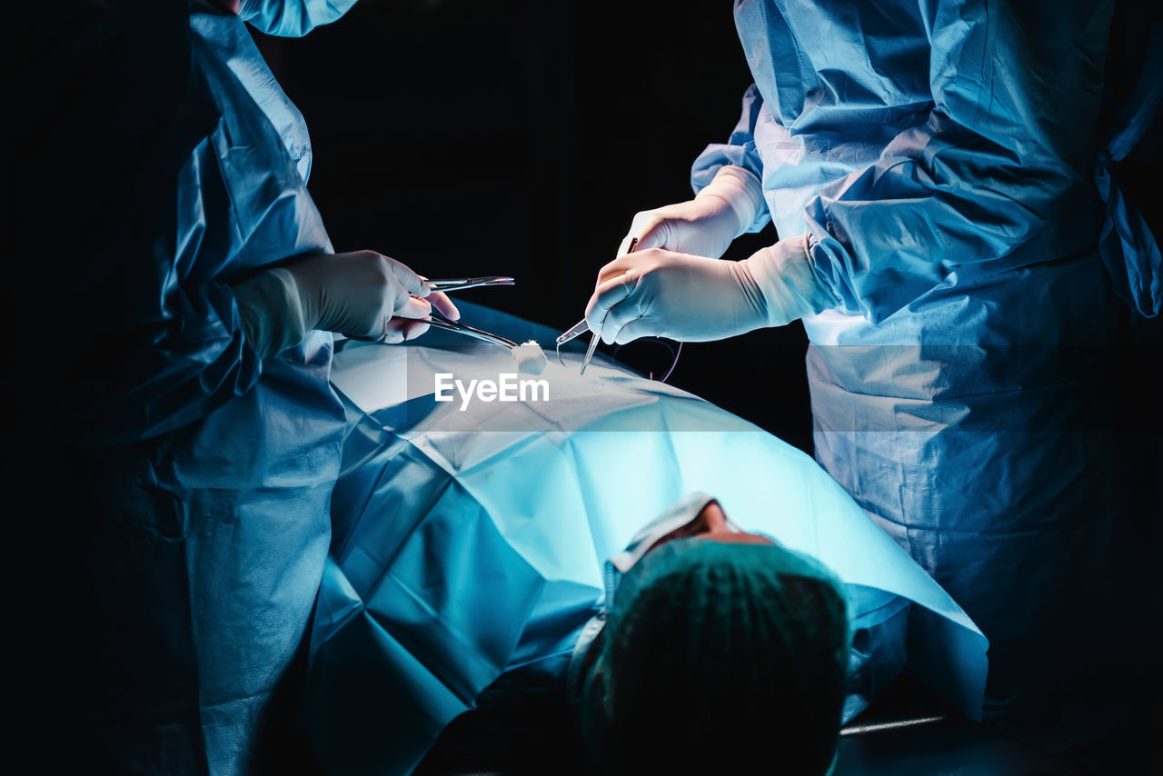 Crop anonymous surgeon and assistant with surgical tools and thread stitching wound of patient during operation in hospital