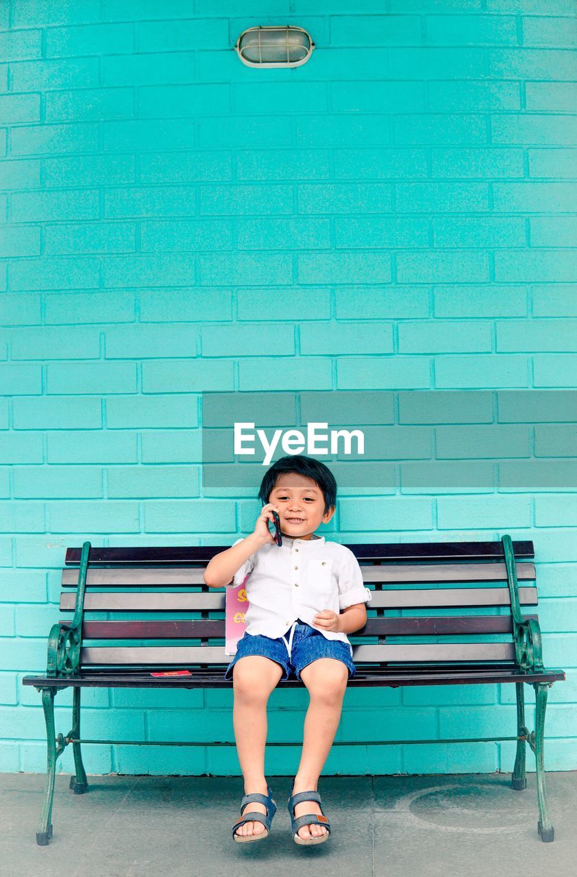 Full length portrait of boy sitting on bench talking on phone against wall