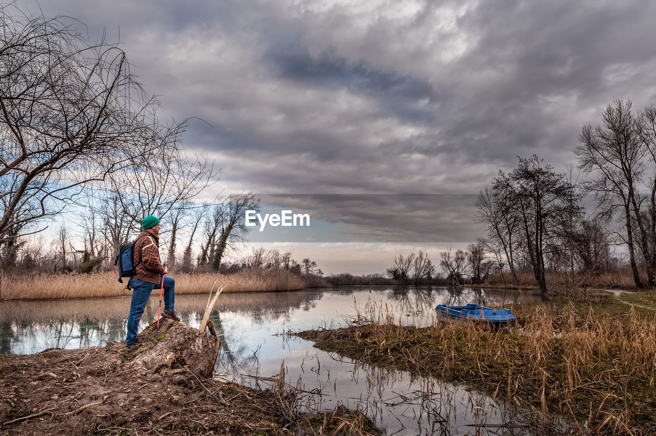 Side view of mature man with backpack standing by lake against cloudy sky