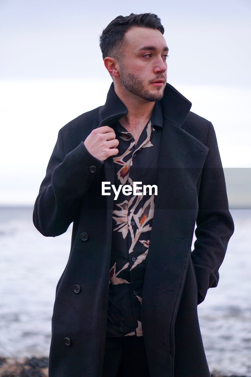 Young man looking away while standing against sea