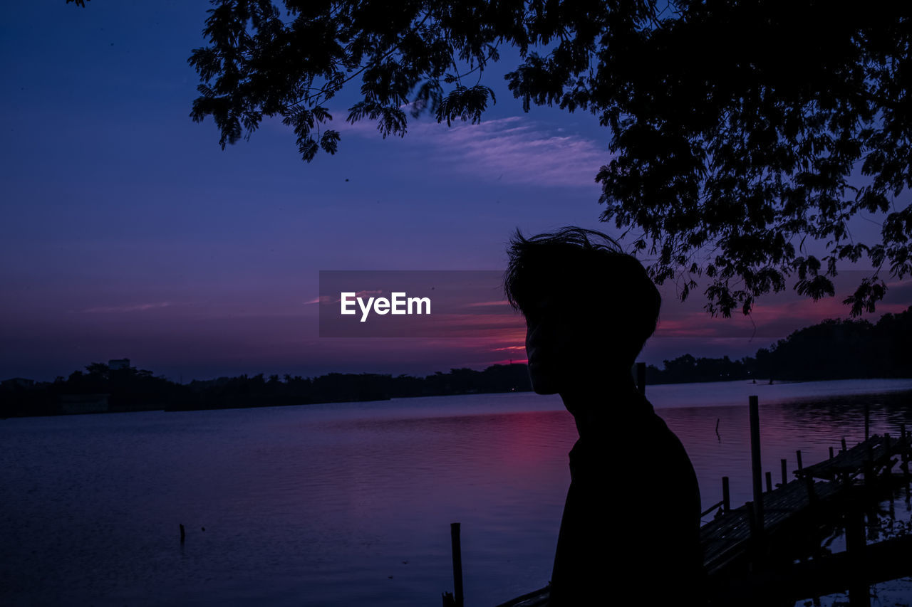 Side view of silhouette man looking at lake against sky during sunset