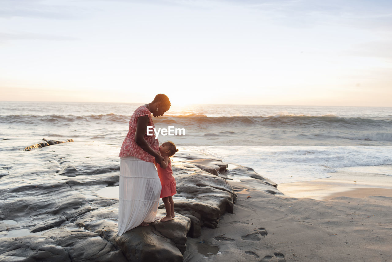 Daughter kissing pregnant mother stomach while standing at beach against sky