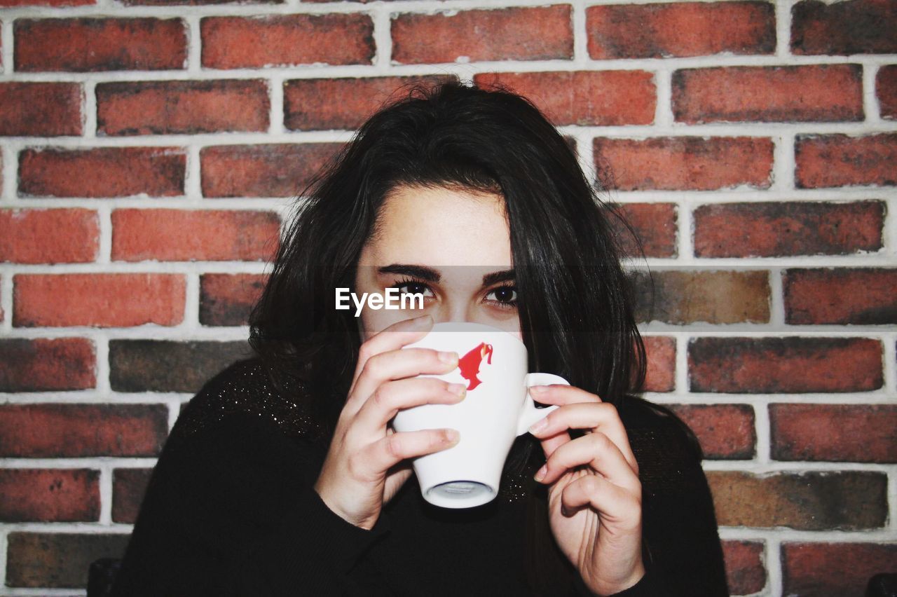 Portrait of young woman drinking coffee against brick wall