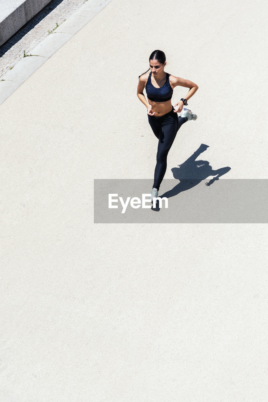 From above of fit female in sportswear running fast along pavement during training on sunny day in city