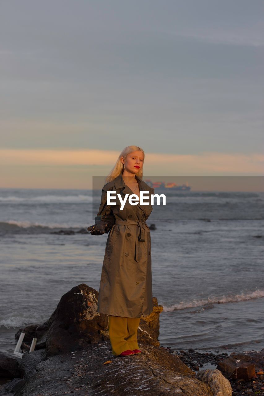 YOUNG WOMAN STANDING ON BEACH AGAINST SKY DURING SUNSET