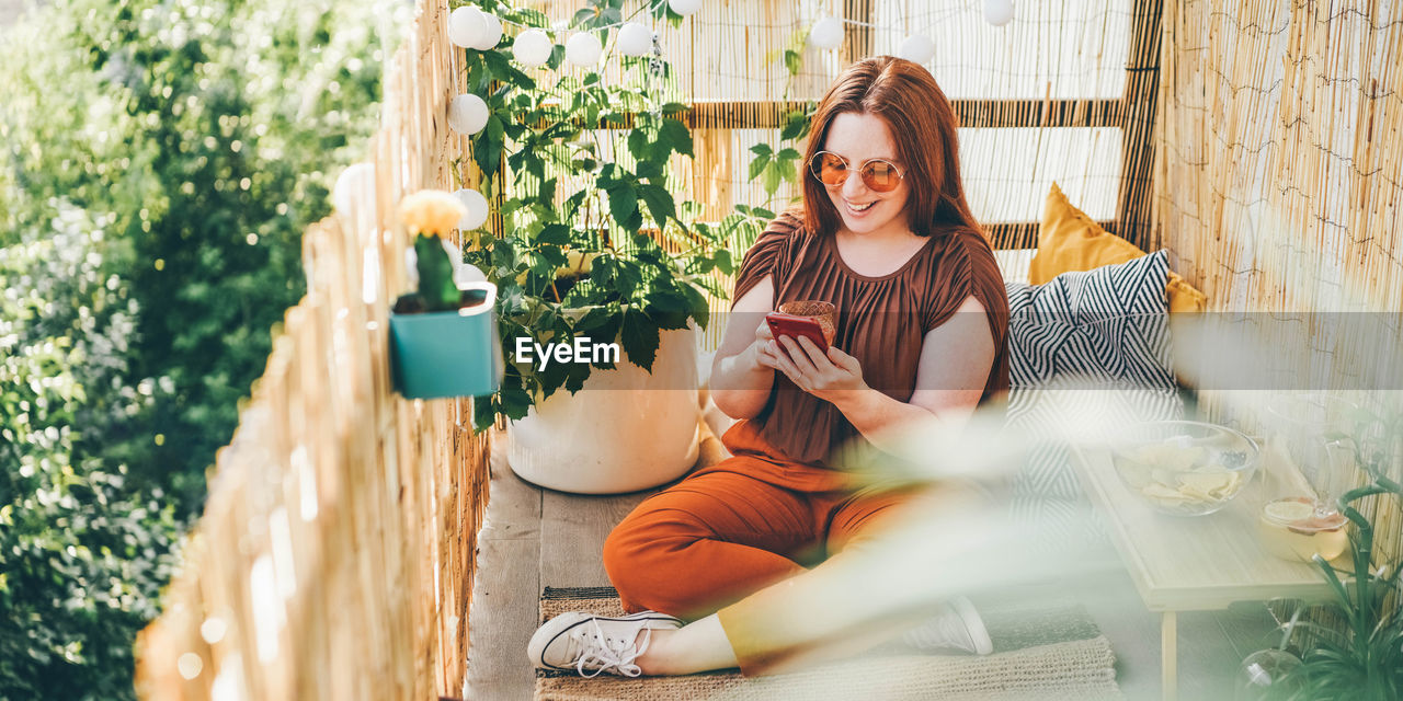 Woman wearing brown blouse and sunglasses works on modern phone siting on pillows on light terrace.