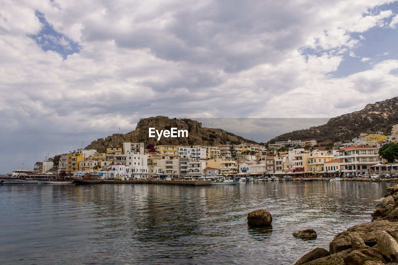 Cityscape by sea against cloudy sky 