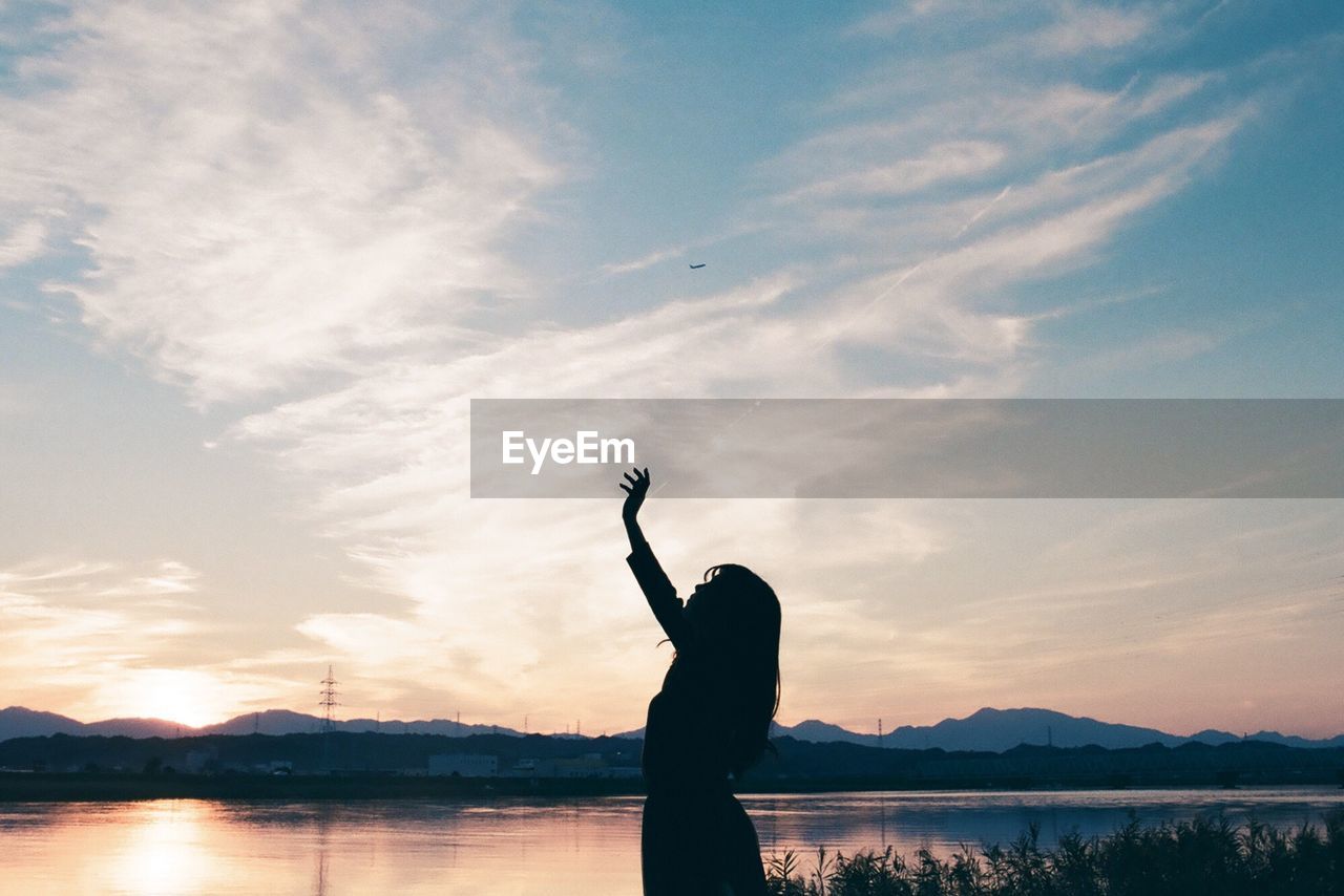 Silhouette woman with hand raised standing by lake against sky during sunset