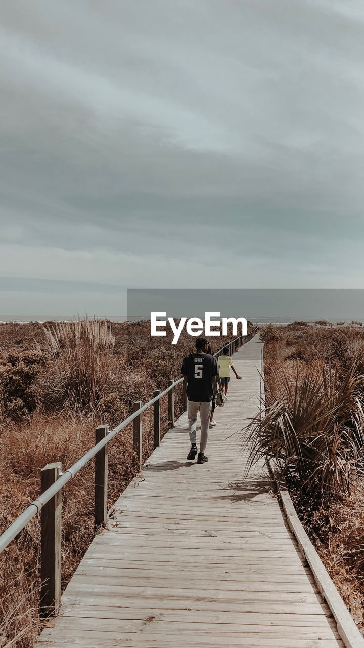 REAR VIEW OF WOMAN WALKING ON FOOTPATH BY RAILING AGAINST SKY