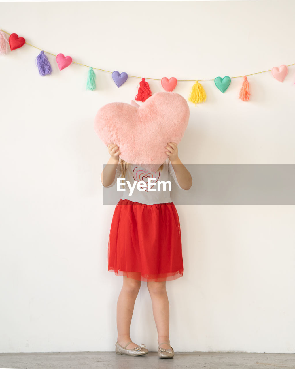 Little girl in red tutu holding pink plush heart pillow over face