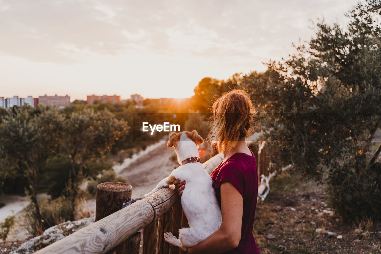 Rear view of woman holding dog against sky during sunset