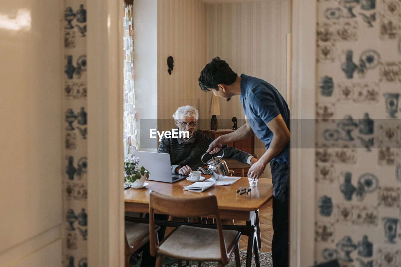 Side view of male healthcare worker serving tea to senior man at home